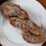 Oblong golden brown twisted donut covered in cinnamon sugar sitting on a white plate.