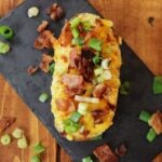 A baked potato with cheese, bacon, and scallions on top of it sitting on a slate board.