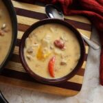 Creamy bowl of soup with peppers and sausage visible in it sitting on a striped cutting board with a spoon.