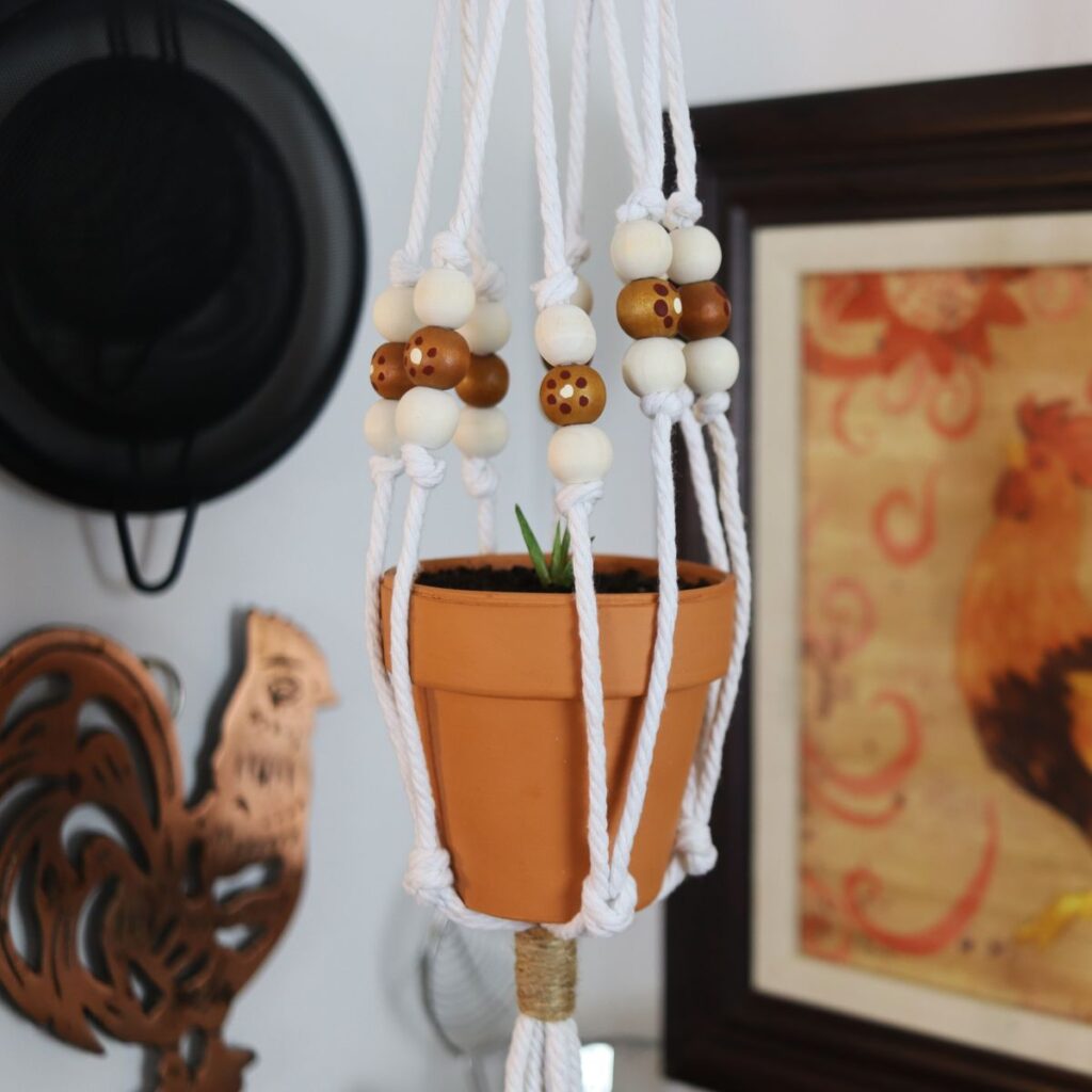 mini macrame plant hanger with painted beads hanging with a picture, trivet, and strainers in the background.