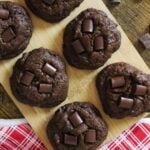 Six double chocolate cookies on a bamboo board next to a red and white checkered cloth.