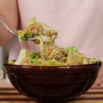 Woman lifting a forkful of out of a bowl of pork, cabbage, carrots, sesame seeds, and scallions.