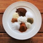 Measured spices and cornstarch placed separately on a white ridged plate over a rustic wood surface.