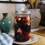 A quart jar with a fermenting lid full of strawberries and blueberries in water sitting on a plate on a counter with kitchen decor visible in the background.