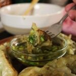 Dipping a piece of a potsticker into a condiment dish of ginger scallion sauce surrounded by other potstickers with a fork. There is a white bowl in the background.