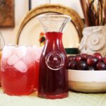 Carafe of grape syrup sitting by an iced purple beverage and a bowl of grapes.