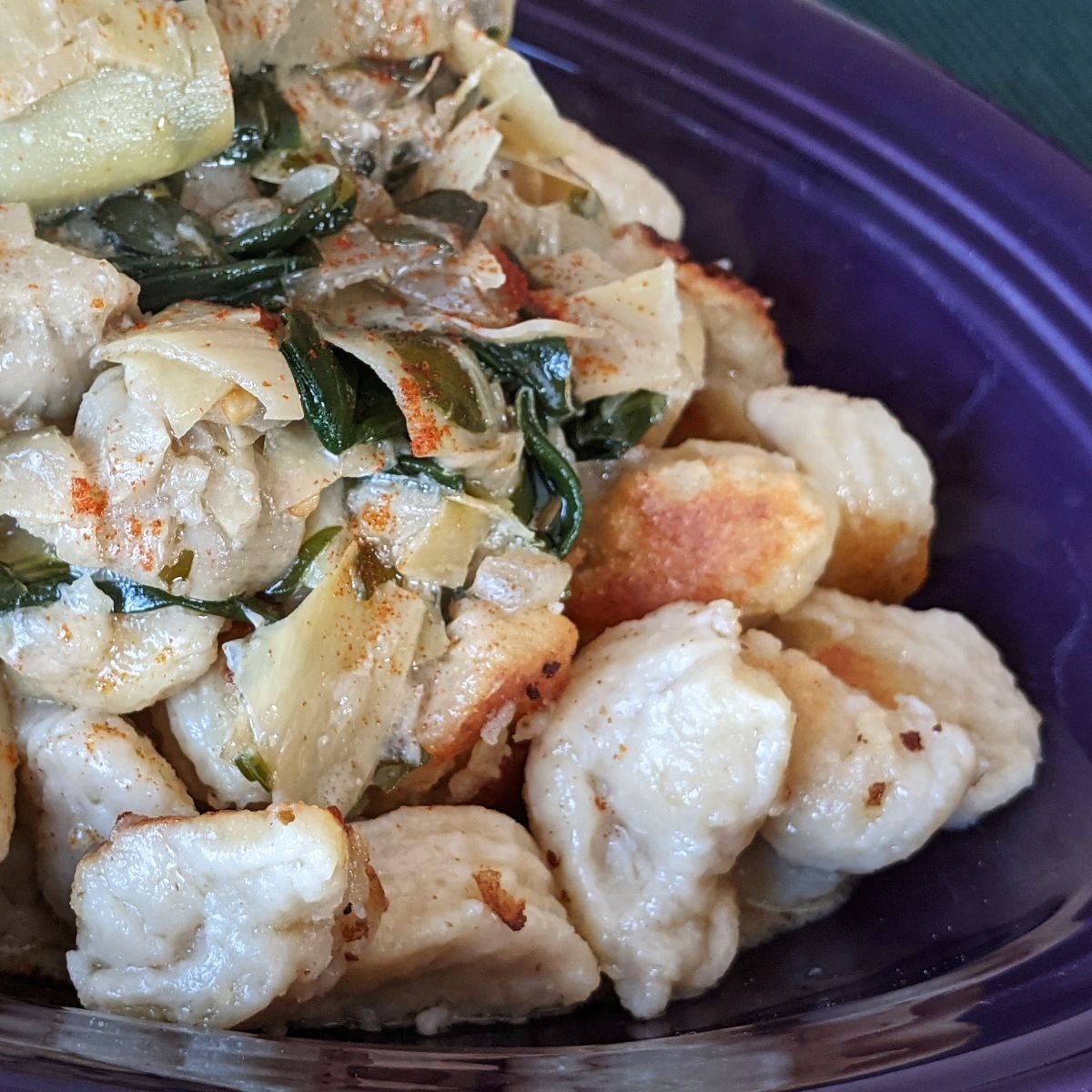 A close-up of golden brown gnocchi topped with a creamy sauce containing artichoke hearts and greens, served in a purple bowl.