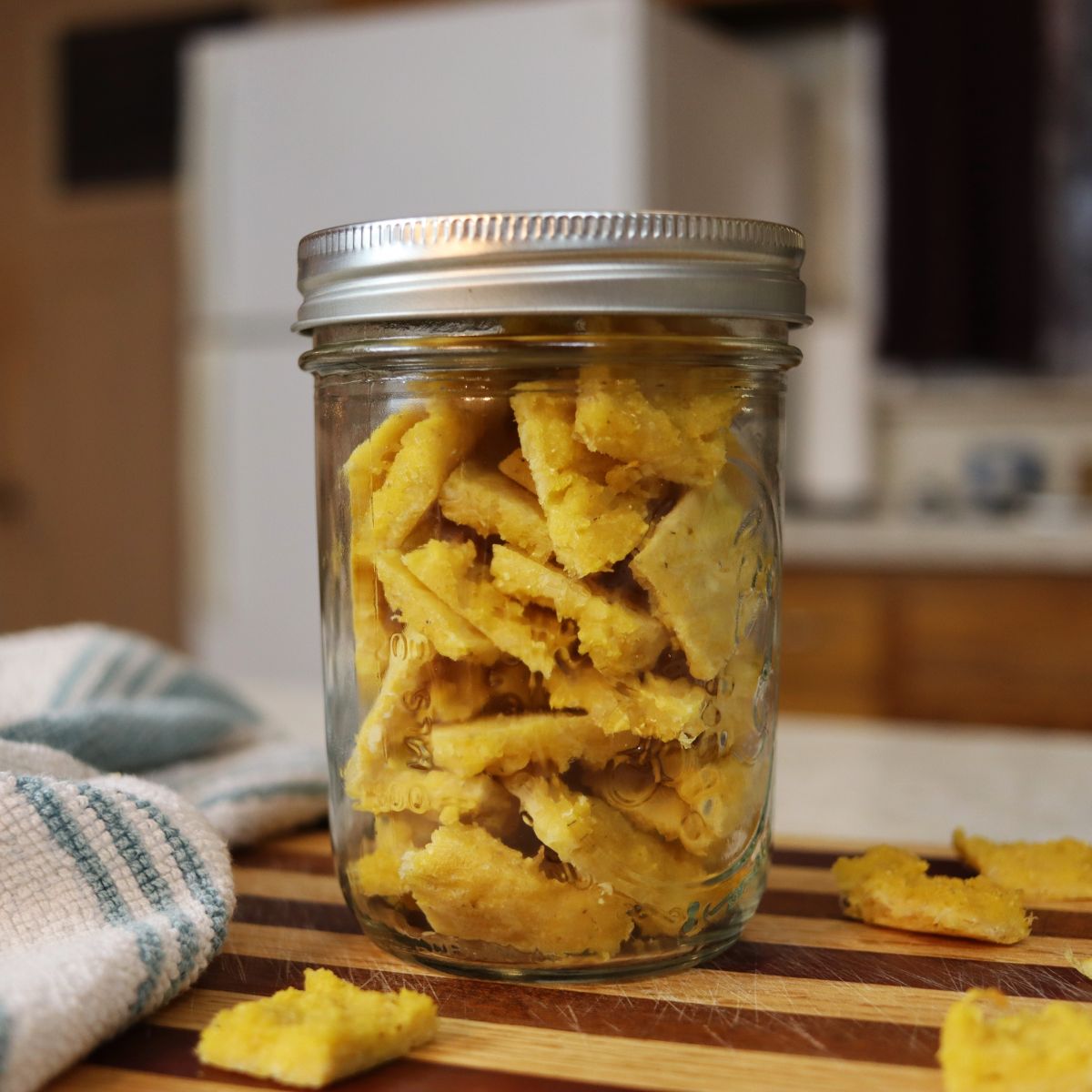mason jar full of homemade square yellow cat treats sitting on a striped board with treats scattered around it.