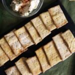 Homemade crackers laid out in two rows on a slate board with a dish of cream cheese sprinkled with paprika visible to the side.