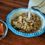 A bowl of bran flakes cereal being topped with oat milk sits on a wooden table.