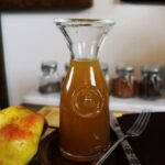 Carafe of golden syrup sitting on a black trivet on a table with pears visible to the side of it.