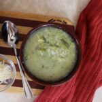 Bowl of broccoli soup on a striped cutting board with a spoon, red cloth, and dish if shredded cheese next to it.