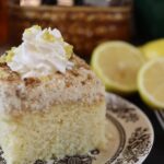 Square piece of yellow cake with crumb topping and whipped cream sitting on a brown and white plate with lemon slices visible in the background.