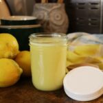 pint mason jar of lemon syrup sitting on a table with a plastic lid, lemons, and a striped cloth sitting next to it.