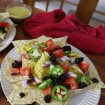 plate of nacho chips topped with vegan cheese, jalapenos, black olives, and tomatoes.