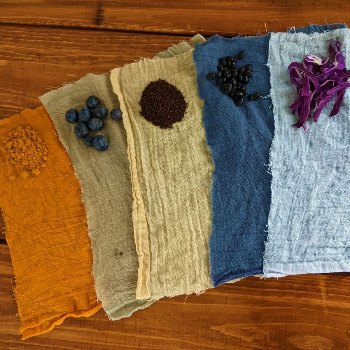 Five pieces of different color fabric laying on a wooden table with a small pile of food in the corner of them.