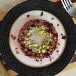 Round cake topped with glaze, rose petals, and chopped pistachios on a plate sitting on a striped cutting board with a knife and fork.