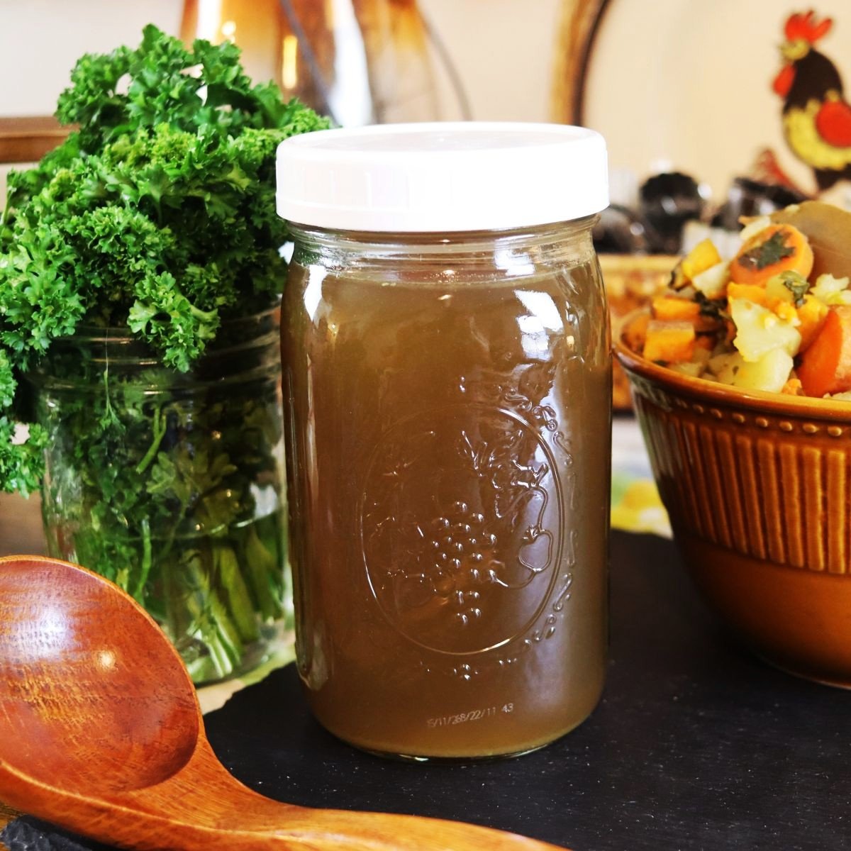 quart jar of broth sitting on a table with parsley and a bowl of vegetables in the background.