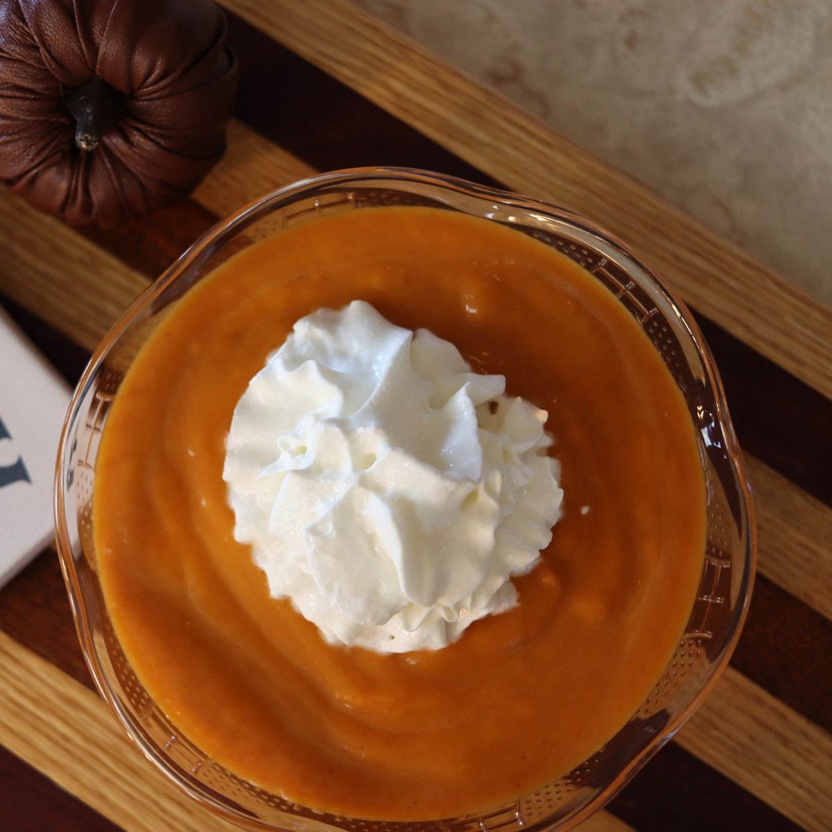 Pumpkin latte for dogs with a dollop of whipped cream on top in a small bowl sitting on a striped cutting board.