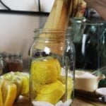 Quart jar of lemons with a fermenting pounder entering the jar with quartered lemons, a dish of salt, and kitchen utensils in the background.