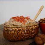 Brown oval dish of sausage dip topped with diced tomatoes with a wooden spoon in it.
