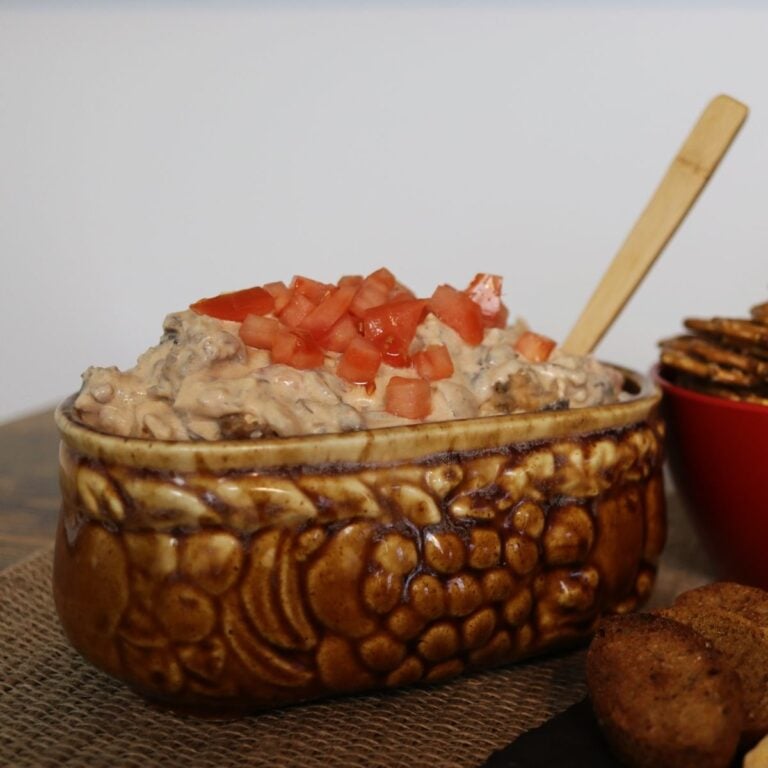 Brown oval dish of sausage dip topped with diced tomatoes with a wooden spoon in it.