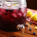 gallon jug with spout full of iced blueberry lemonade on a table.