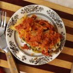 Stuffed cabbage roll on a plate topped with tomato sauce and saurkraut sitting on a striped cutting board with a knife and fork to the side of it.