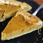 Slice of pumpkin galette being held above a dark brown surface with a serving utensil with more slices in the background.