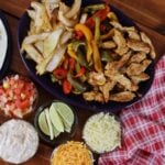 Platter of sliced chicken with bell peppers and onions, bowls of salsa, lime wedges, shredded cheese, and sour cream on table.