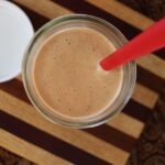 A creamy, orange sauce in a glass jar seen from above, with a red spatula inserted, placed on a striped wooden surface.