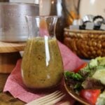 Glass carafe of golden dressing with herbs sitting on a red napkin next to a salad.