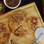 Golden vegetable pancake cut in quarters on a cutting board with another pancake to the side, a pizza cutter, and a bowl of sauce and a bowl of scallions on the side.
