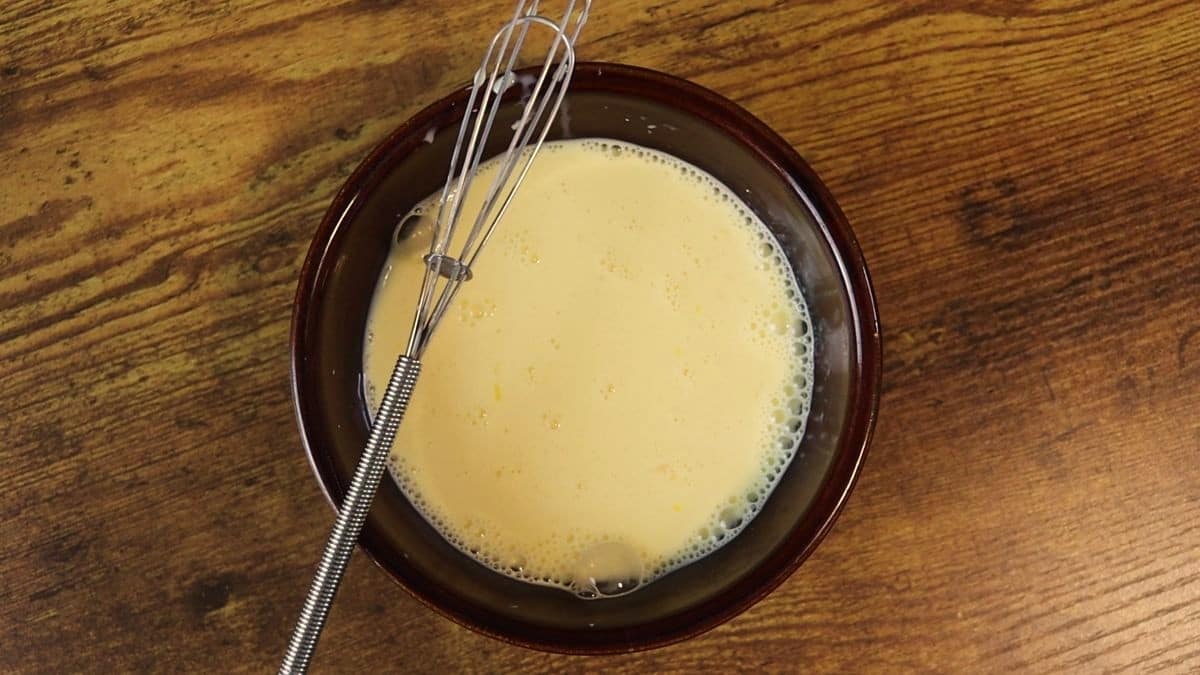 Creamy yellow mixture in a bowl with a small whisk resting on it's edge sitting on a wooden surface.