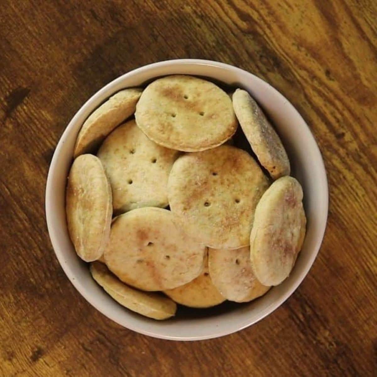 White bowl full of round golden brown crackers sitting on a wooden table.