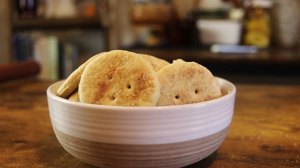 A white ceramic bowl filled with round, golden-brown crackers sits on a wooden table, with a blurred kitchen background.