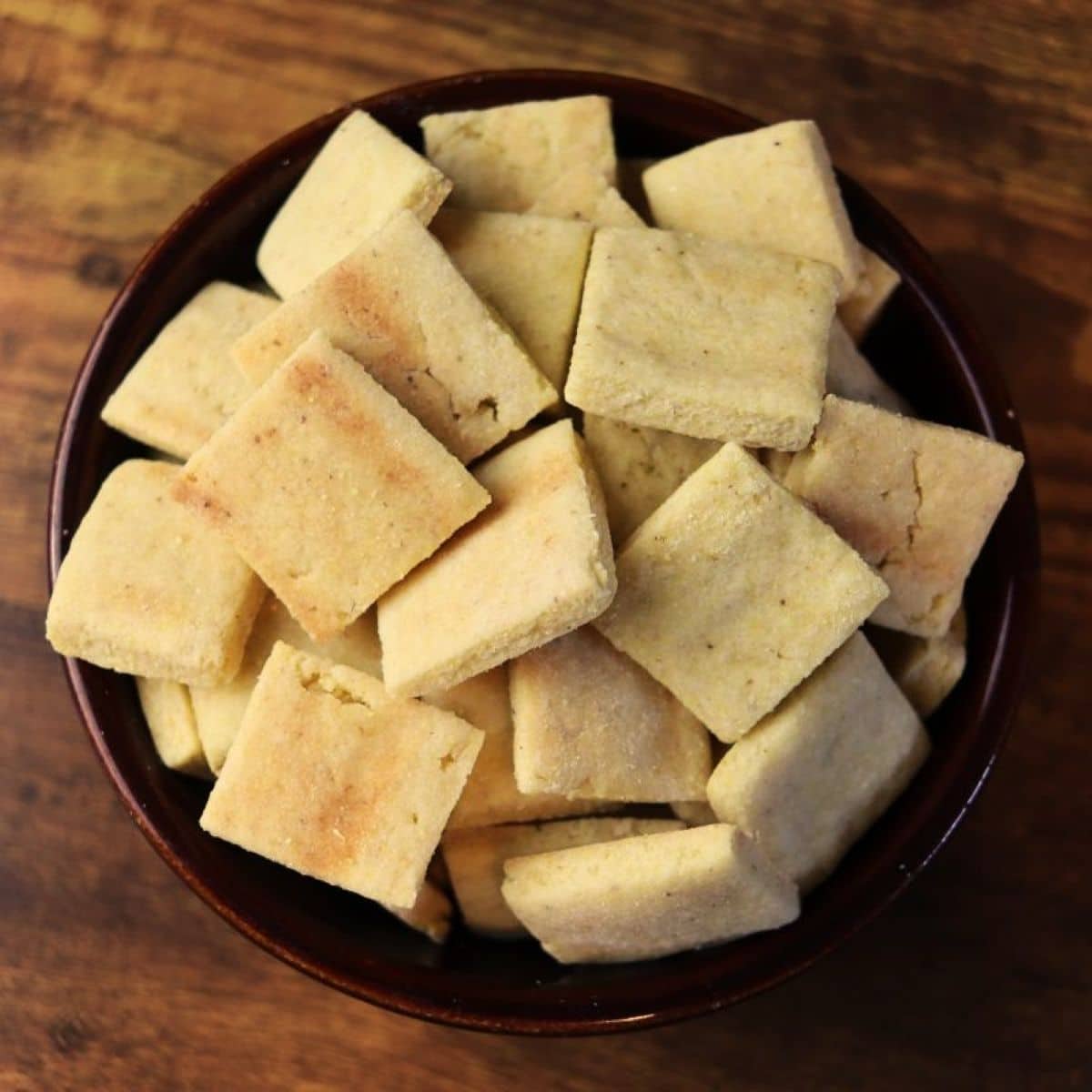 Bowl of square golden colored crackers on a wooden surface.