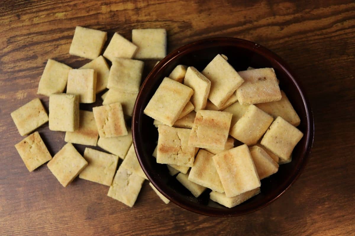 A brown bowl filled with square, golden-brown crackers sits on a wooden surface. More crackers are scattered around the bowl, showing their baked texture.