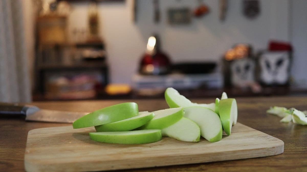 Slices of green apple on a cutting board on a counter with a knife and apple cores on the counter behind it and a kitchen counter with tools and a small stovetop blurred but visible in the background.
