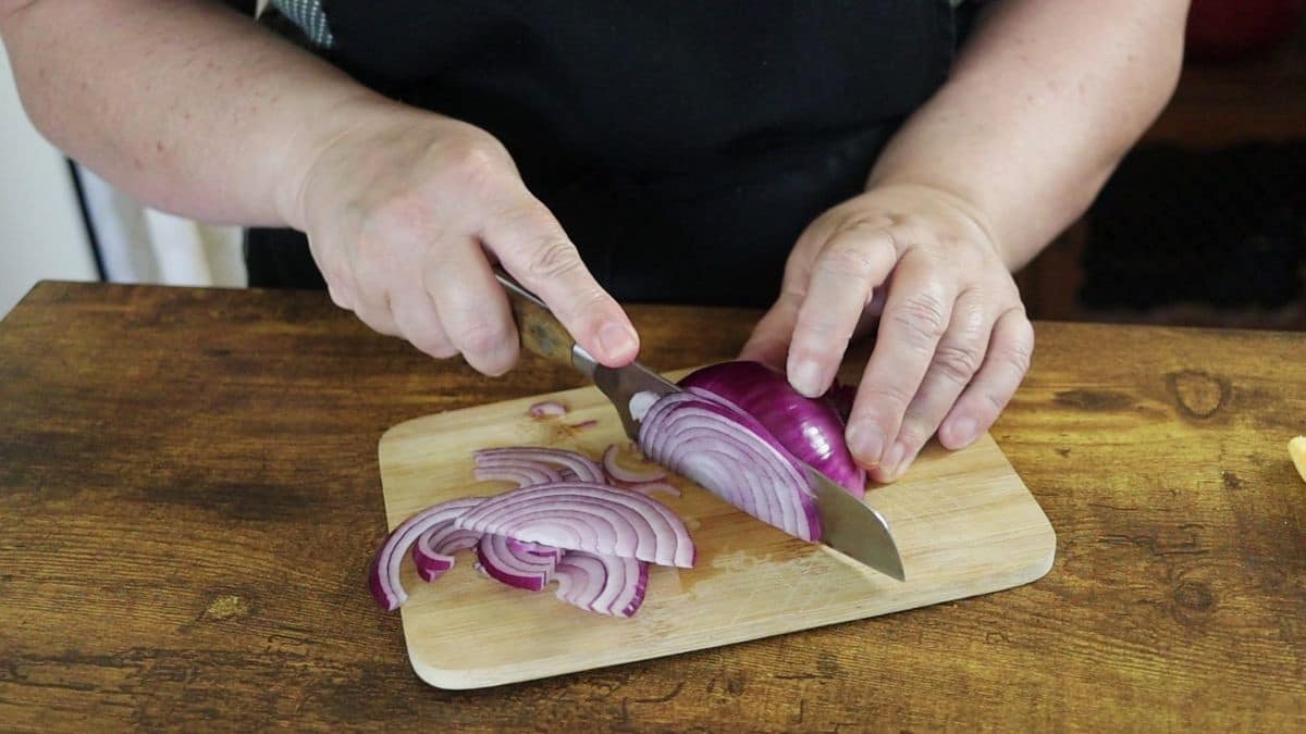 Woman's hands cutting an onion on a small cutting board sitting on a wooden countertop with knife.