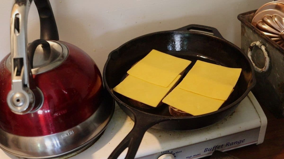 Two slices of bread topped with yellow cheese in a cast iron skillet on a small stovetop with the edge of a tea kettle showing next to it.