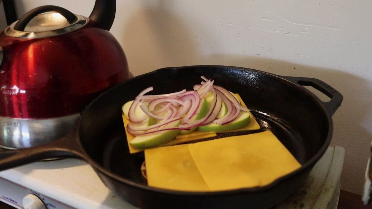 Two slices of bread topped with yellow cheese, one with apple and onion slices on it too, in a cast iron skillet.