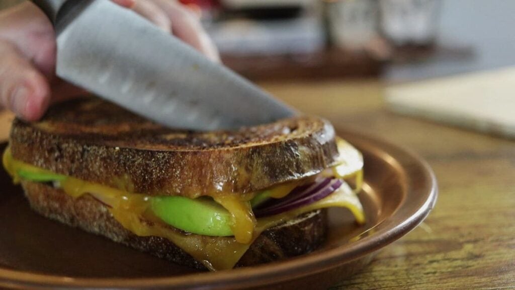 Grilled sandwich on dark bread with yellow cheese, green apple, and red onion visible in it sitting on a brown plate being cut by a knife.