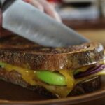 Knife cutting through a grilled sandwich with melted cheese, green apple, and red onion inside.