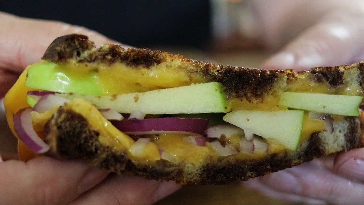 Close-up of hands holding a grilled cheese sandwich filled with melted cheddar, green apple slices, and red onion on toasted bread.