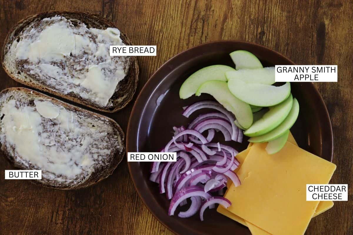 Two slices of buttered rye bread on a wooden surface next to a brown plate holding sliced Granny Smith apple, red onion, and cheddar cheese, each ingredient clearly labeled.