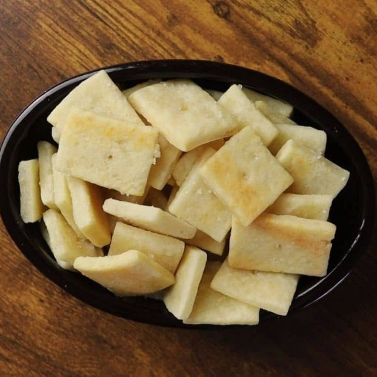 Dark colored oval dish full of small square crackers sitting on a wooden surface.