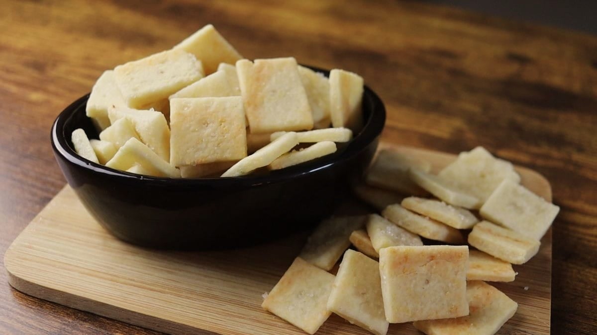 A black bowl filled with square, homemade cheese crackers sits on a wooden cutting board, with more crackers scattered beside the bowl, all on a wooden table.
