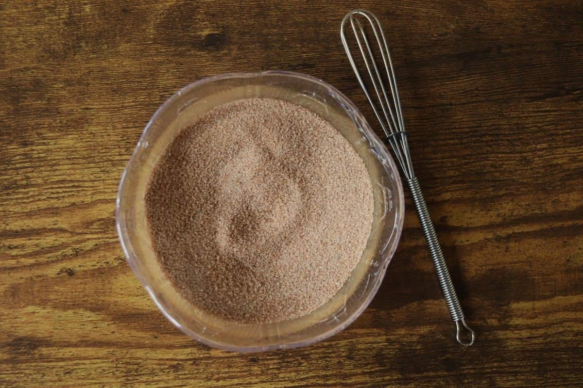 Bowl of cinnamon sugar sitting next to a whisk on a wooden surface.