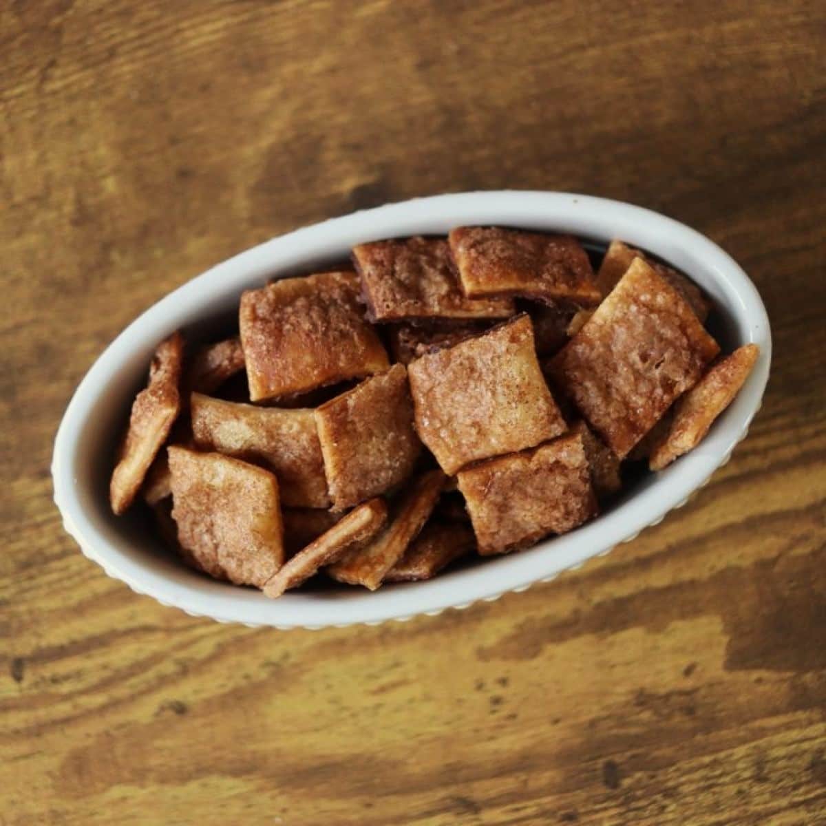 White oval dish full of brown cinnamon sugar coated crackers sitting on a wooden surface.